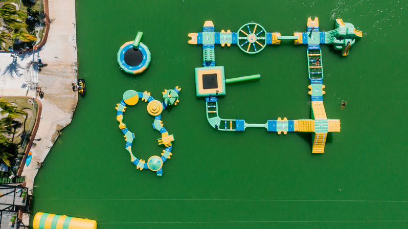Aerial view of a colorful inflatable water park obstacle course on green lagoon water beside a palm-lined tropical shoreline, with slides, trampolines and a jet ski.