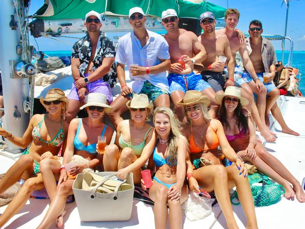 Sunlit catamaran party: group of friends in colorful swimsuits and sun hats enjoying drinks on deck with turquoise tropical ocean and clear blue sky.