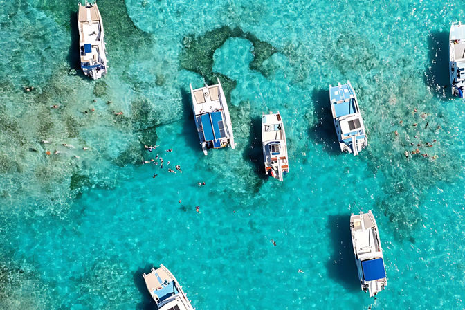 Aerial view of a bright turquoise lagoon dotted with white boats anchored over shallow coral reef and snorkelers swimming in crystal-clear water