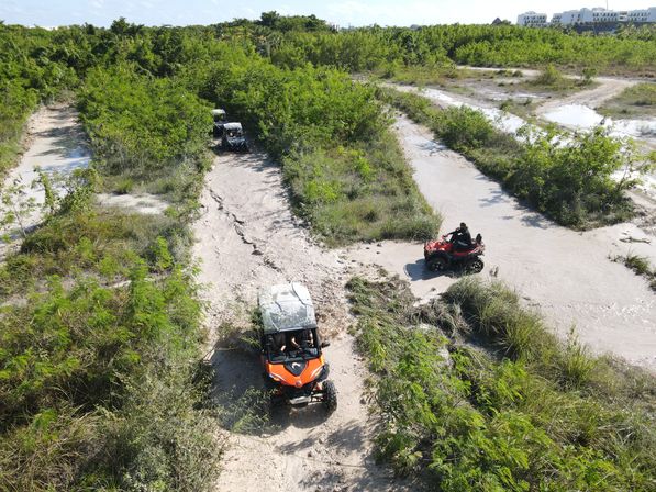 Aerial drone view of an off-road adventure: an orange UTV leading a convoy of UTVs and a red ATV navigating muddy sandy trails through dense green scrubland under a sunny sky.