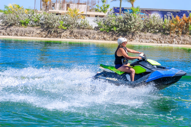 Person in a white helmet riding a neon green-and-blue jet ski across clear coastal waters, creating splashes with a sunlit tropical shoreline in the background.