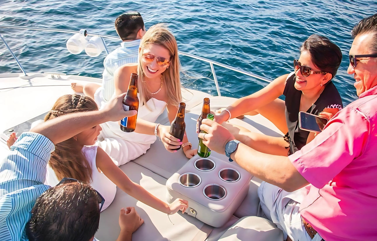 Friends clinking beer bottles on a sunlit yacht deck with blue ocean in the background — casual boat party on the water.