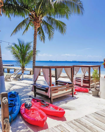 Tropical beachfront with swaying palm trees, wooden cabanas draped in white curtains, red and blue kayaks on white sand, lounge chairs and a pier stretching into the clear blue sea under a sunny sky.