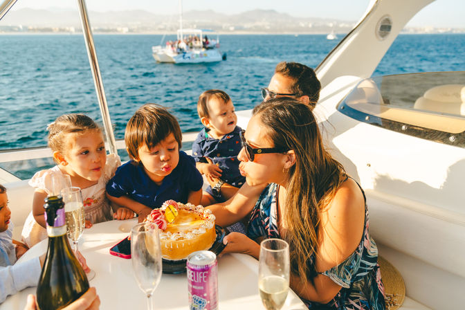 Sunlit family birthday on a yacht — kids and adults gathered around a cake blowing out candles on a boat with sparkling ocean and distant shoreline in the background.
