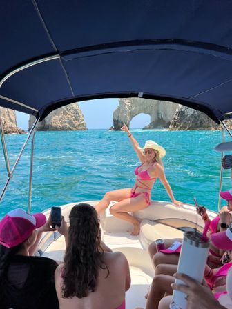 Group on a boat photographing a woman in a pink bikini and straw hat posing by a dramatic natural sea arch over turquoise water on a sunny boat tour.