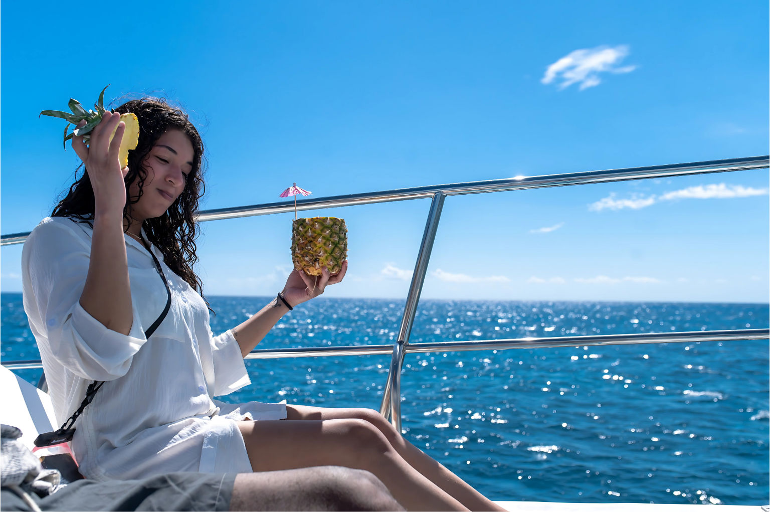 Woman in a white cover-up relaxing on a boat deck, holding a pineapple cocktail with a mini umbrella against a sparkling blue ocean and clear sunny sky.