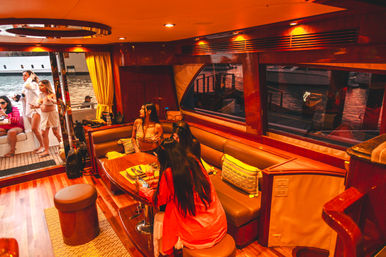 Sunlit luxury yacht interior at a marina — warm wood-paneled salon with cushioned banquette and table, a group of women in swimsuits socializing by an open door with docked boats visible outside.