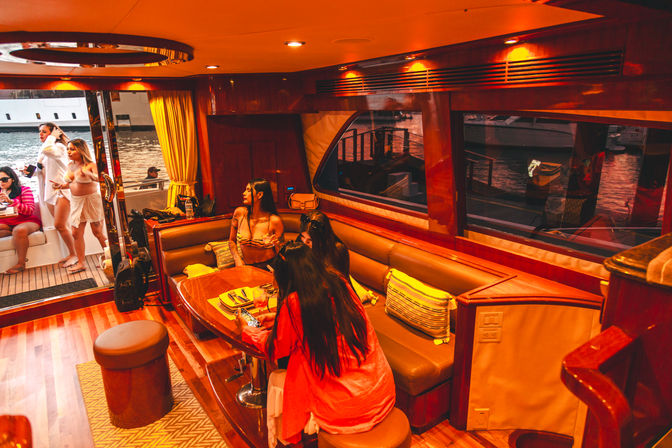 Sunlit luxury yacht interior at a marina — warm wood-paneled salon with cushioned banquette and table, a group of women in swimsuits socializing by an open door with docked boats visible outside.