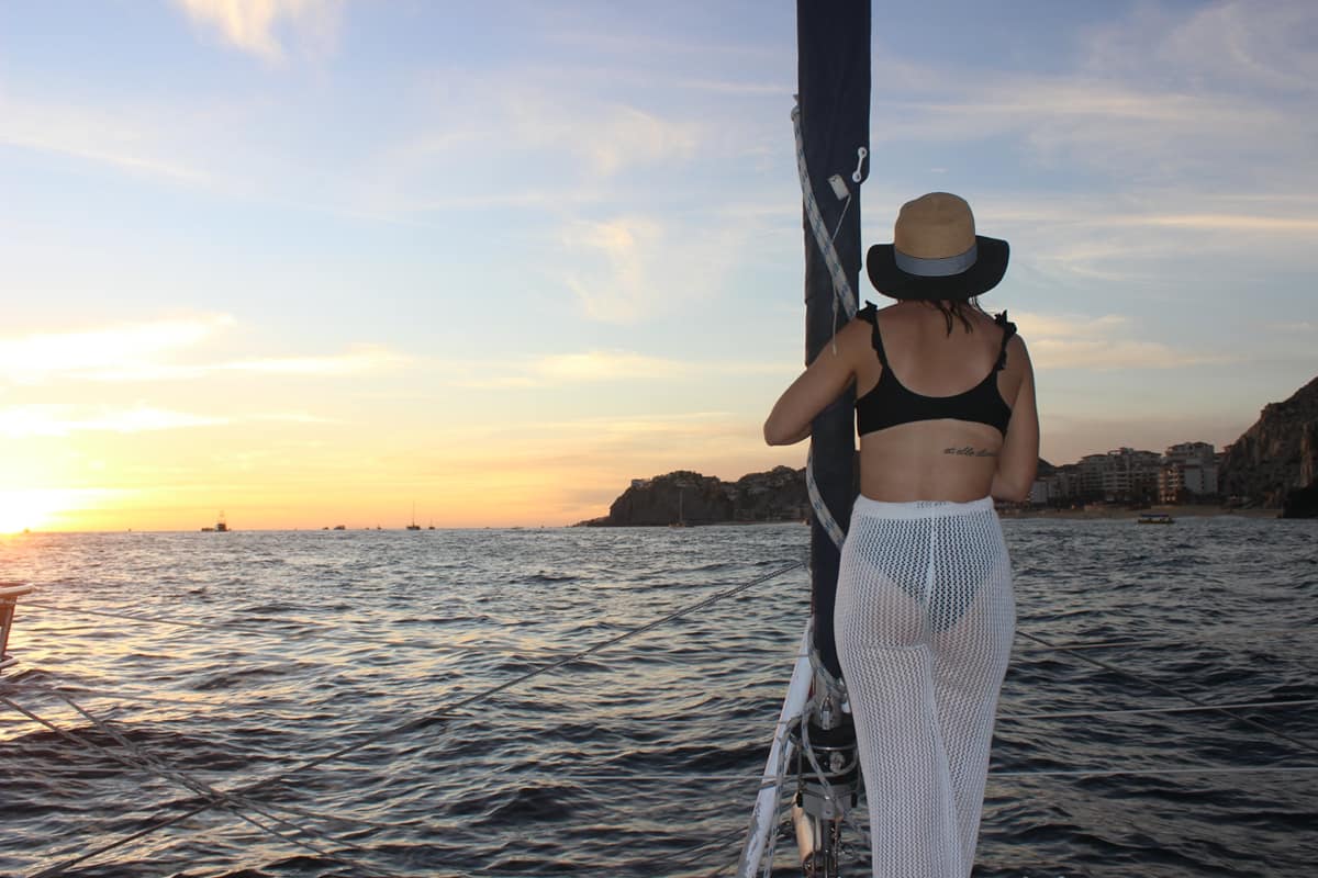 Person in a sun hat and black bikini top stands on a sailboat holding the mast, enjoying a golden sunset over the ocean with rocky coastline and boats on the horizon.