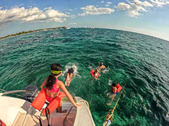 Children and an adult jumping from a boat into clear turquoise tropical water near a shoreline, wearing orange life jackets and snorkeling gear with sailboats on the horizon.