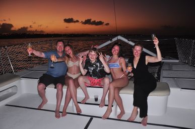 Smiling group of five in swimwear holding drinks and posing on a catamaran deck at a vibrant orange sunset over calm tropical waters — festive sunset boat cruise scene.