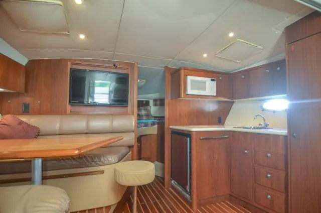 Cozy yacht cabin interior with warm wood paneling, compact galley (microwave, sink, mini-fridge), leather dinette and table, flat-screen TV and sleeping berth in the background
