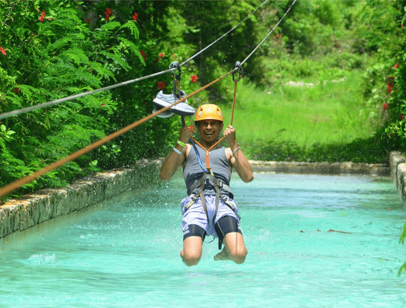 Person wearing a helmet and harness ziplining low over a turquoise pool in a lush tropical setting, shoes clipped to the cable and water splashing beneath.