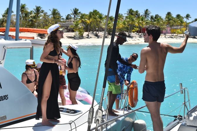 Young adults wearing captain hats at a sunny catamaran boat party in turquoise tropical waters off a palm‑fringed island beach.