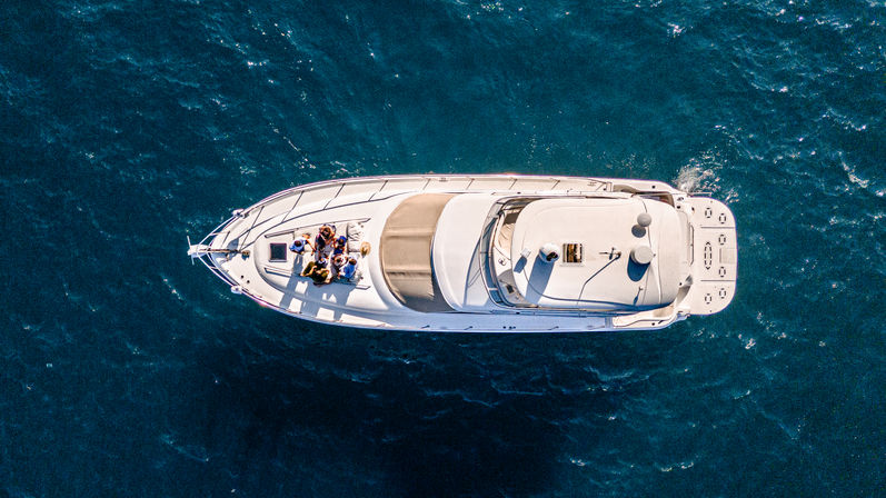 Aerial drone shot of a white luxury yacht on deep blue open sea, with a small group relaxing on the sunlit bow and deck details visible.