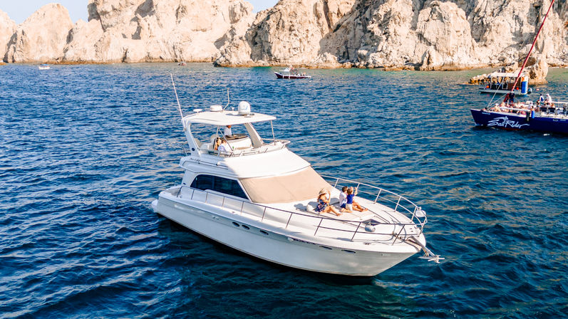 White luxury yacht with sunbathers on the bow cruising on deep blue water near sunlit rocky coastal cliffs, with nearby tour boats on a sunny vacation day