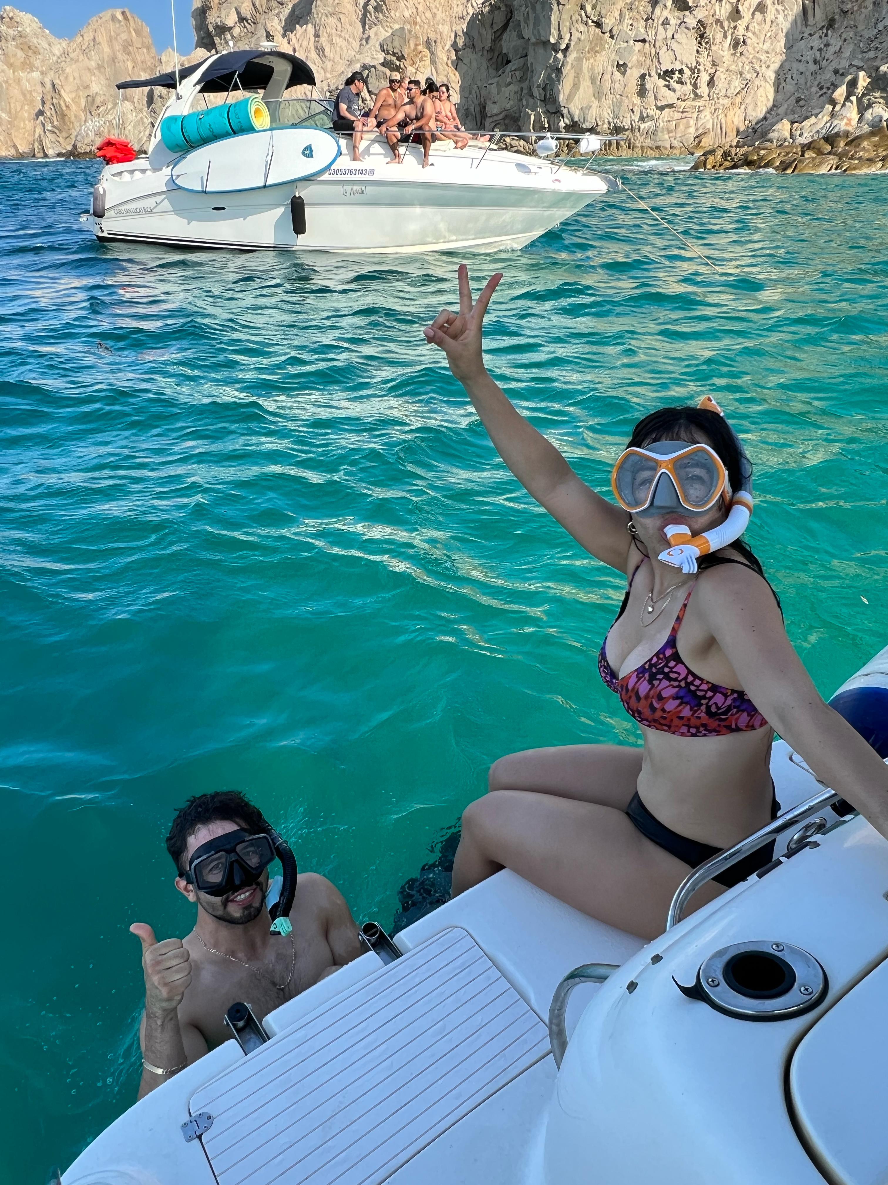 Two snorkelers—one in the water giving a thumbs-up and one on the boat flashing a peace sign—next to a small white motorboat anchored in turquoise coastal waters with rocky cliffs in the background.