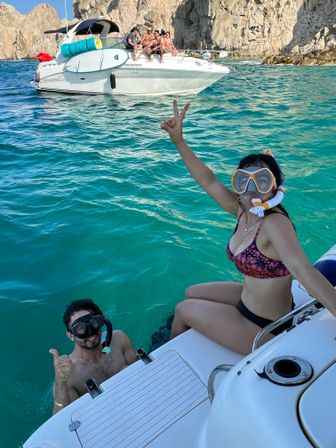 Two snorkelers—one in the water giving a thumbs-up and one on the boat flashing a peace sign—next to a small white motorboat anchored in turquoise coastal waters with rocky cliffs in the background.