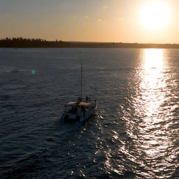 Small boat cruising on the ocean at sunset, golden sunlight forming a glittering path across rippling water toward a low tree-lined coastline.