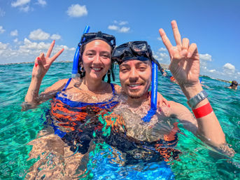 Happy snorkelers — a smiling couple wearing black masks and blue snorkels flashing peace signs while floating in clear turquoise ocean water under a sunny blue sky, vibrant tropical vacation scene.
