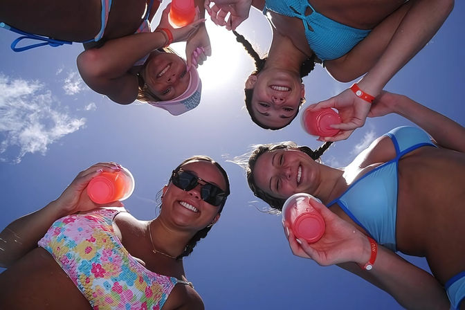 Overhead shot of four smiling women in colorful bikinis holding pink drinks up to the camera under a bright blue summer sky, summertime beach/pool vibe.