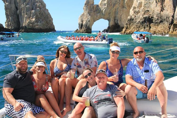 Smiling group of adults on a sunny Cabo San Lucas boat tour near the El Arco rock arch, turquoise water and tour boats in the background, some holding drinks.