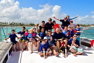 Cheerful multigenerational group on a catamaran deck during a family boat trip in turquoise tropical waters with a palm-lined shoreline and blue sky