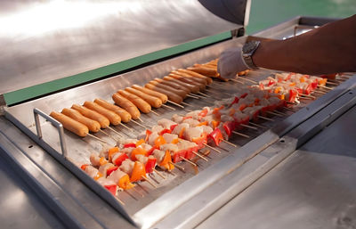 Stainless-steel grill with rows of skewered hot dogs and colorful chicken-and-bell-pepper kebabs being arranged by a gloved hand at an outdoor food stall
