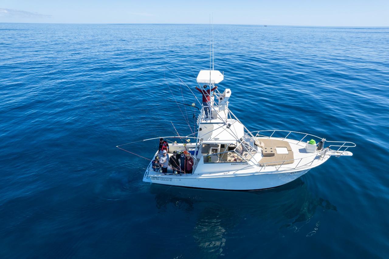 Aerial view of a white sportfishing boat with anglers and multiple fishing rods in calm, deep-blue offshore waters under a clear sky.