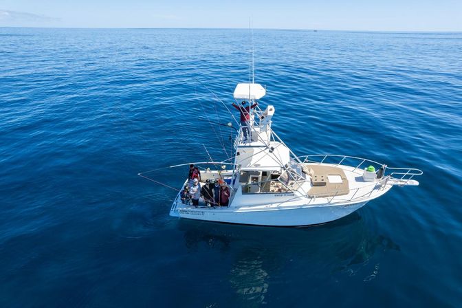 Aerial view of a white sportfishing boat with anglers and multiple fishing rods in calm, deep-blue offshore waters under a clear sky.