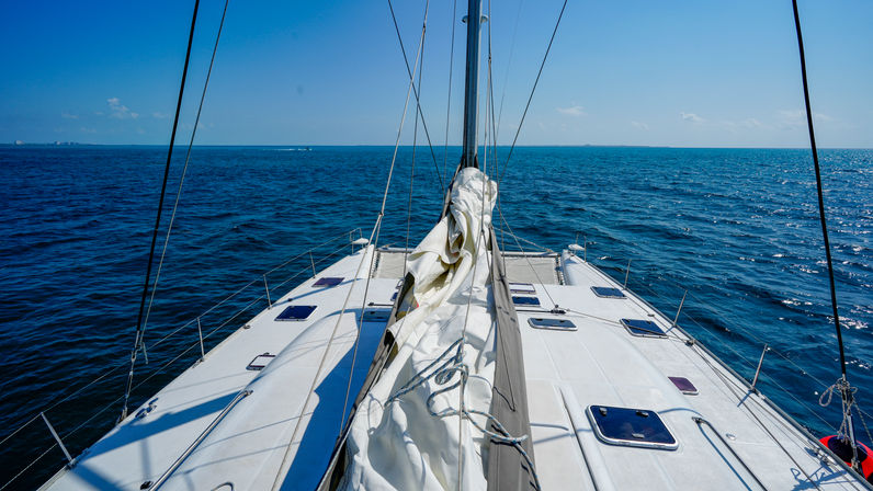 Sunlit sailboat bow on the open blue sea, deck with a rolled white sail, rigging lines and small hatches under a clear blue sky on the horizon.