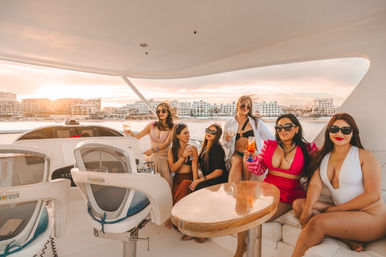 Group of women friends in swimwear sipping cocktails on a luxury yacht at sunset with waterfront resort buildings in the background, enjoying a sunset cruise.