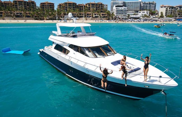 Aerial view of a white-and-navy luxury yacht anchored in turquoise water off a sandy beachfront resort, with four women in swimsuits sunbathing and jumping from the bow and a blue floating platform nearby.