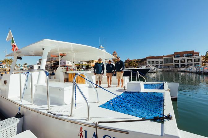 Three crew members on the deck of a white catamaran with blue netted bow, docked at a sunny marina/harbor with waterfront buildings and a clear blue sky.