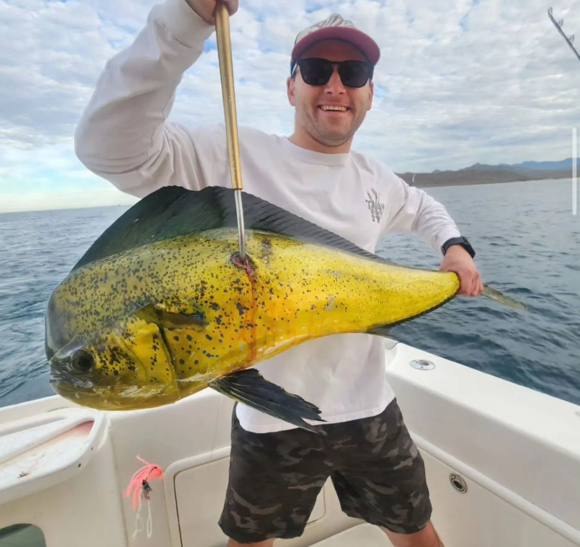 Smiling angler on a sportfishing boat holding a large yellow-green mahi-mahi (dolphinfish) over offshore coastal waters with distant mountains and cloudy sky.