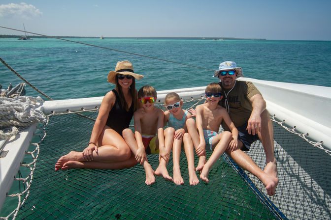 Two adults and three children relaxing on a catamaran trampoline net, wearing swimsuits, sunglasses and sun hats, smiling with turquoise tropical ocean and distant shoreline in the background.