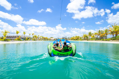 Two people water-tubing on a bright green inflatable across a clear turquoise lagoon toward a palm-lined tropical beach under a sunny blue sky