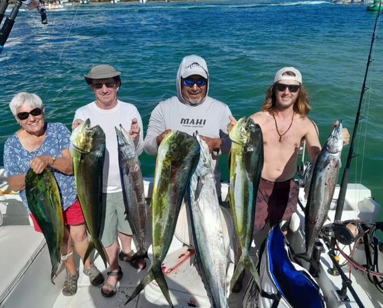 Four anglers on a sportfishing boat proudly holding a mixed catch of large green-gold mahi‑mahi (dorado) and silver wahoo with turquoise coastal waters and sunny sky in the background.