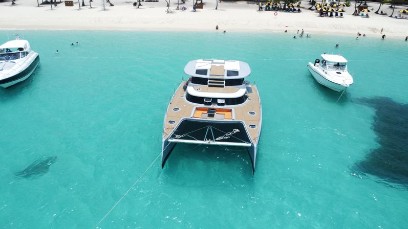 Aerial view of a luxury catamaran anchored in clear turquoise water off a sun-drenched white-sand tropical beach, flanked by two motorboats and swimmers