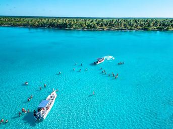 Aerial view of a sunny tropical lagoon with vivid turquoise water, two boats and groups of people wading in shallow crystal-clear water near a palm-lined shoreline.