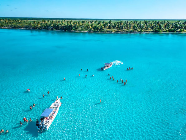 Aerial view of a sunny tropical lagoon with vivid turquoise water, two boats and groups of people wading in shallow crystal-clear water near a palm-lined shoreline.