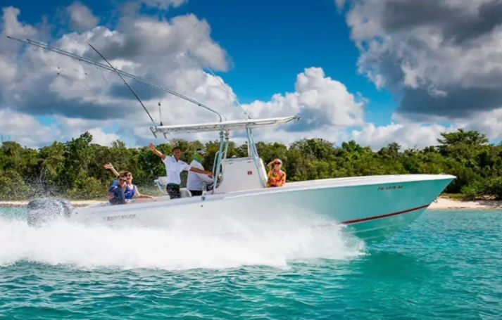 Center-console speedboat zipping through clear turquoise water past a tropical island shoreline, passengers waving under a bright blue sky with puffy clouds.