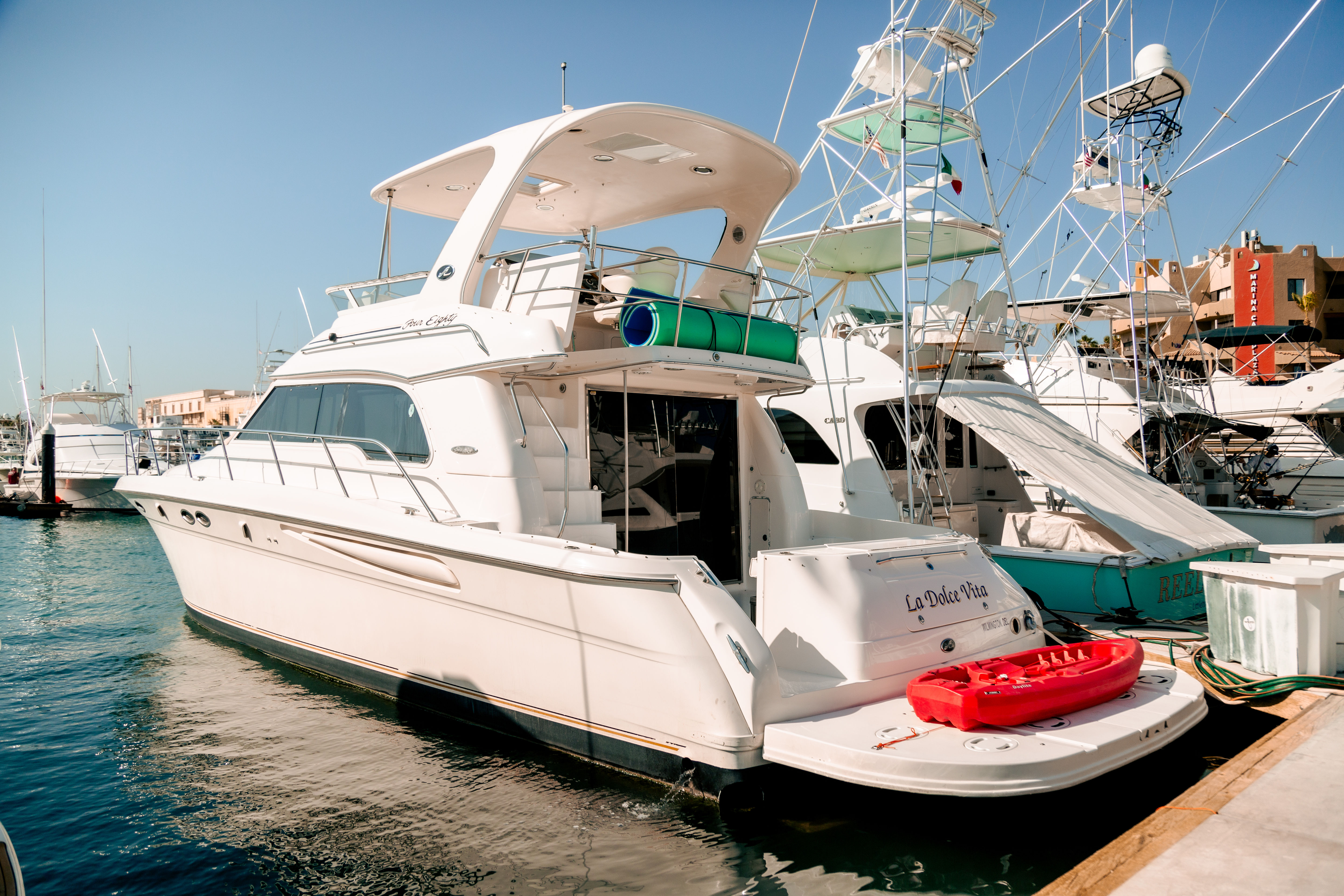 White luxury motor yacht docked in a sunny coastal marina, stern platform holding a bright red kayak with neighboring sportfishing boats and clear blue sky