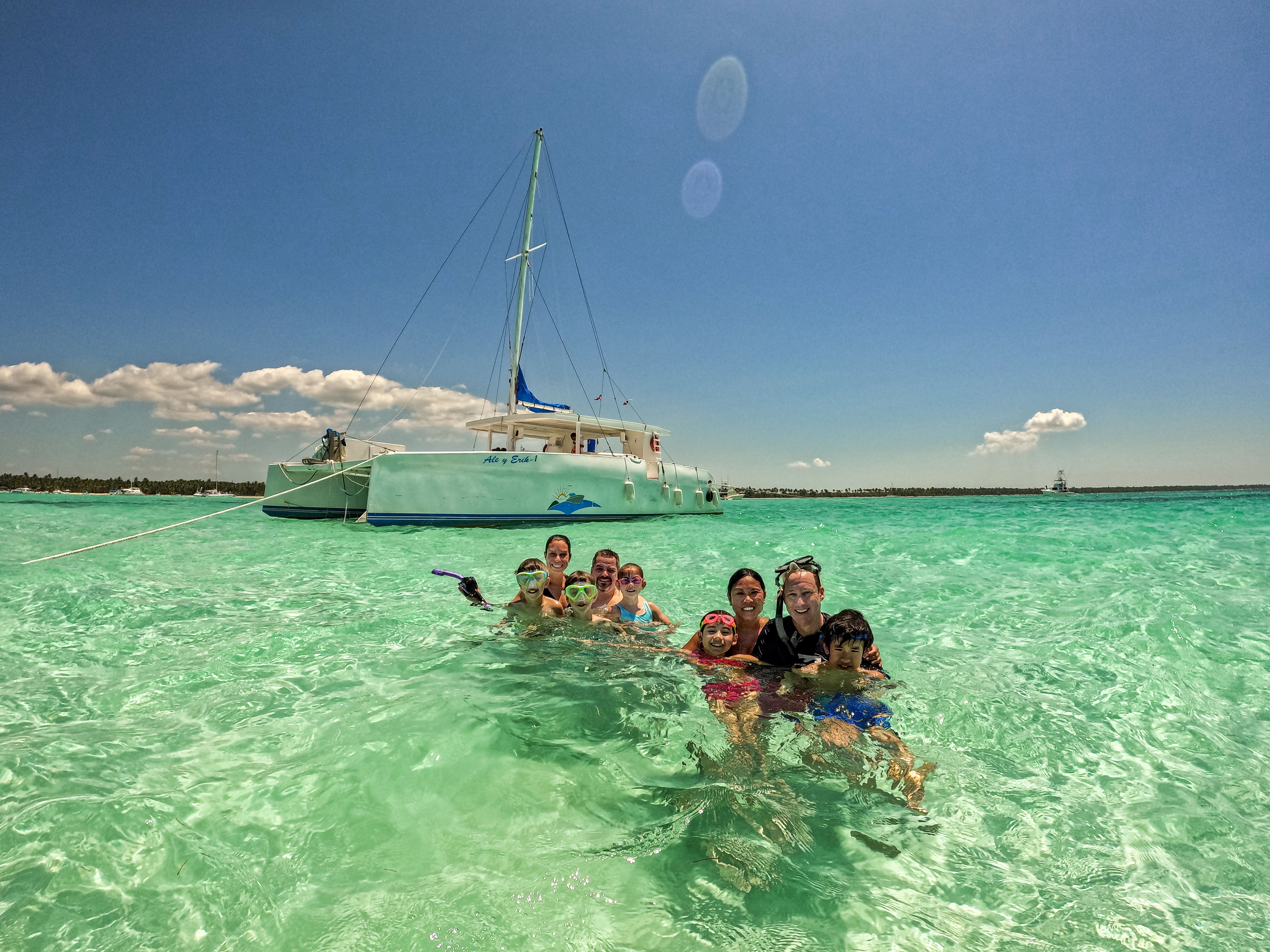Smiling group of swimmers and children wearing snorkel masks in crystal-clear turquoise water beside an anchored white catamaran under a bright blue tropical sky