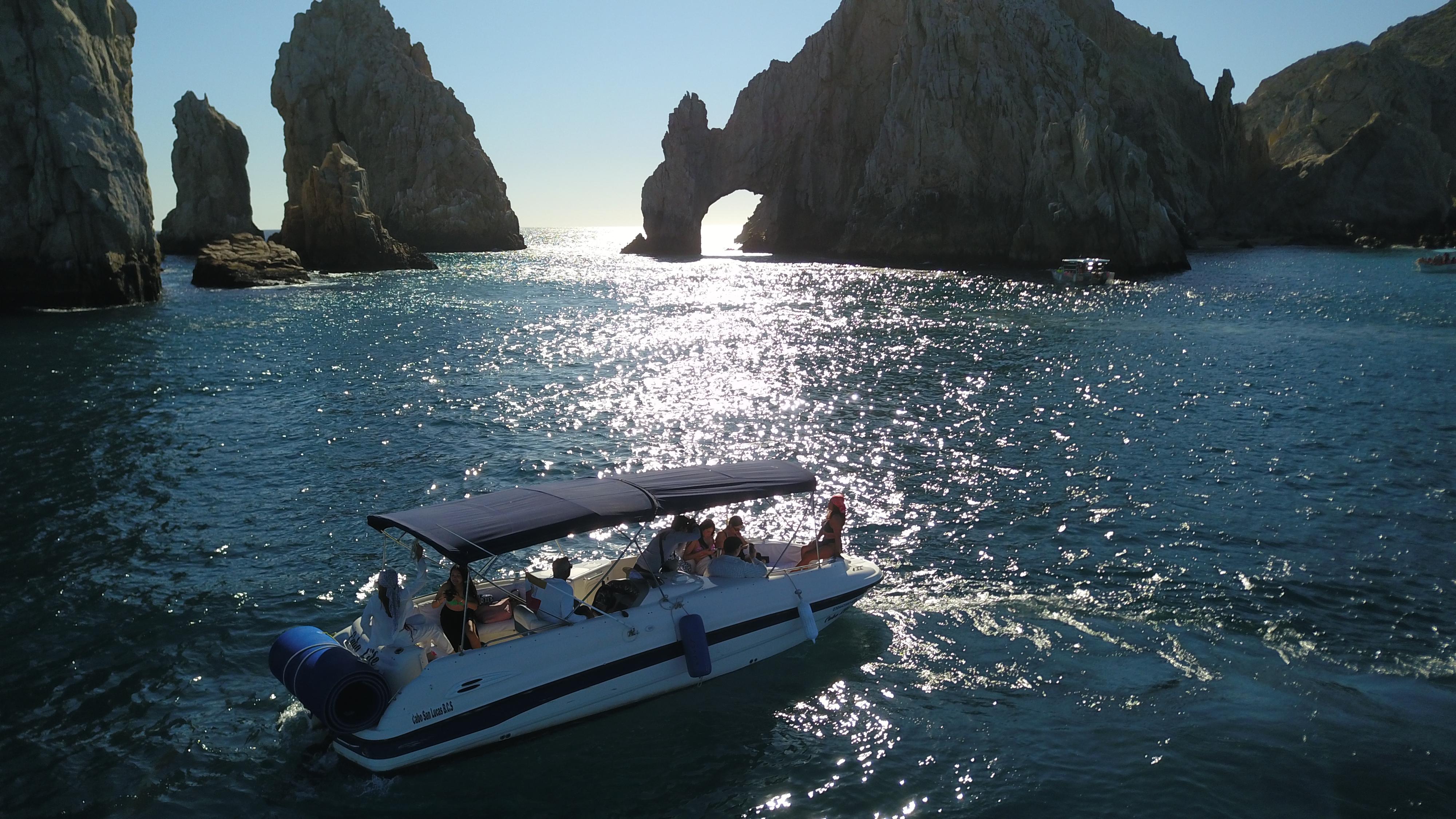 Motorboat with passengers cruising on sparkling blue water toward the sunlit natural rock arch at Cabo San Lucas, with rugged sea stacks in the background.