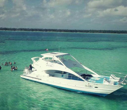 White luxury yacht anchored in crystal-clear turquoise tropical sea near a sandy shoreline, with people swimming and snorkeling beside the boat under a partly cloudy sky