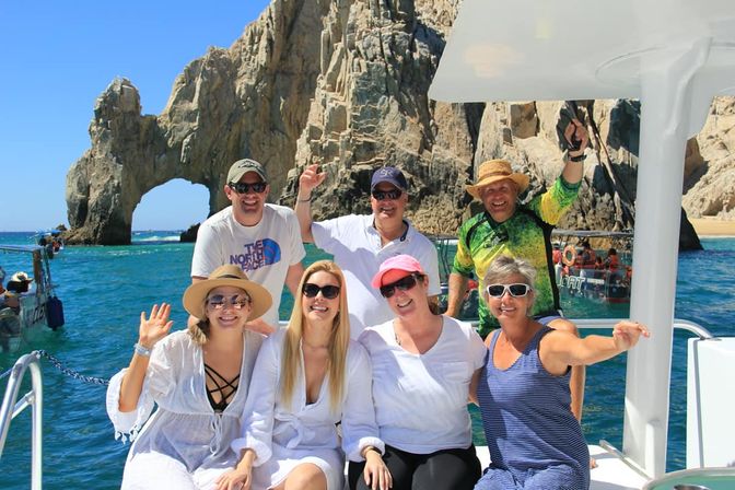 Cheerful group of seven adults on a boat waving and smiling in turquoise water with the iconic rock arch of Cabo San Lucas and a bright blue sky in the background.