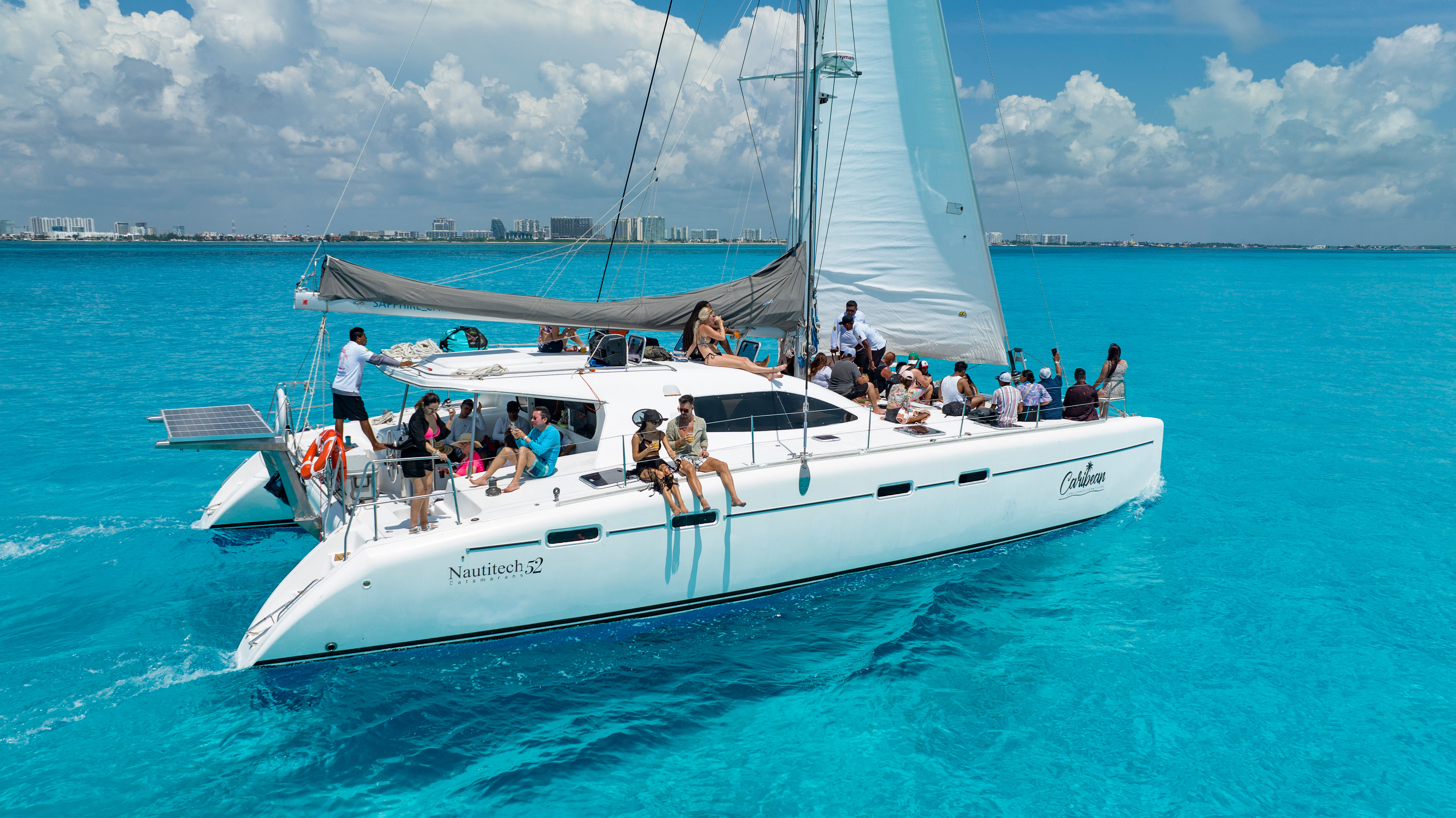 White catamaran cruise with people lounging and socializing on deck, sails up, gliding across clear turquoise Caribbean waters under a sunny blue sky with a distant coastal skyline.