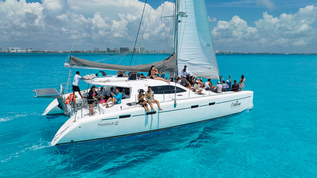 White catamaran cruise with people lounging and socializing on deck, sails up, gliding across clear turquoise Caribbean waters under a sunny blue sky with a distant coastal skyline.