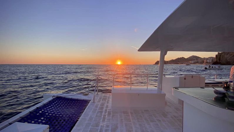 Golden sunset over the ocean seen from a white boat deck with a blue netted trampoline, stainless railings and shaded canopy, sun reflecting on rippling water and a rocky coastline with boats on the horizon.